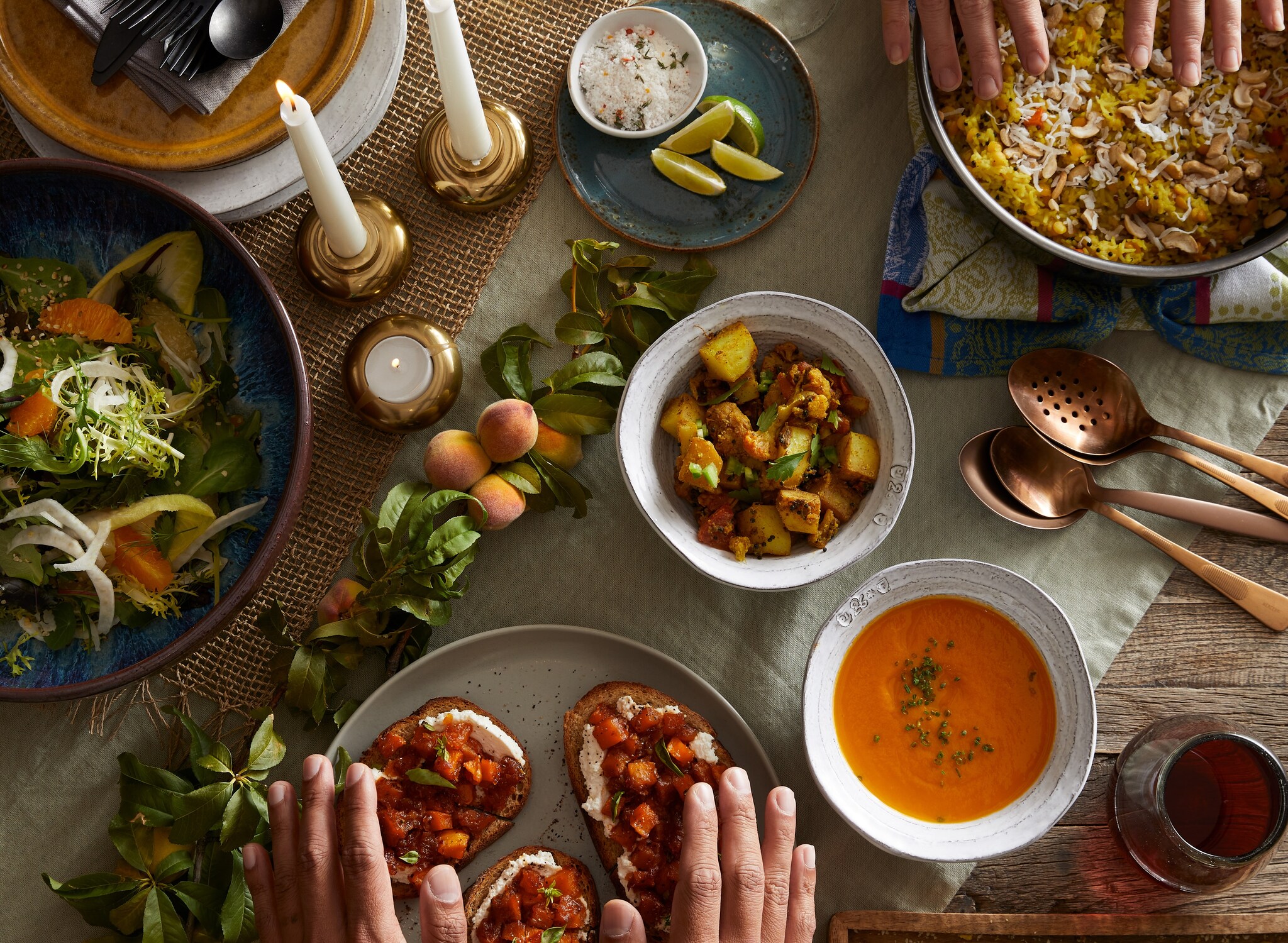 A rustic table is set with candles, brass spoons, a branch of peaches, and plates of colorful food, including roast potatoes and tomato soup.