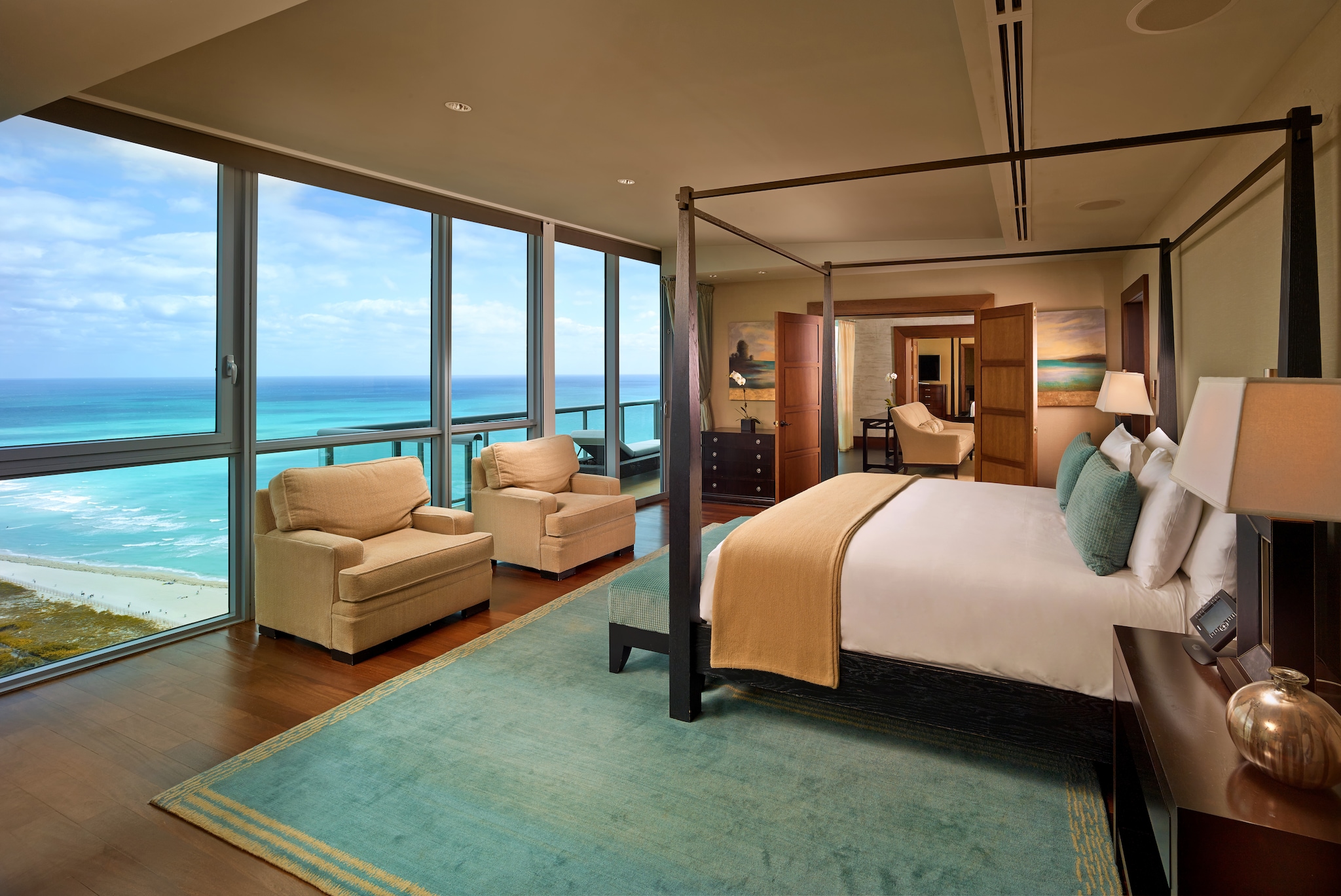 In a suite, a big bed faces a row of floor-to-ceiling windows. Doors on one end of the room open into a living space.