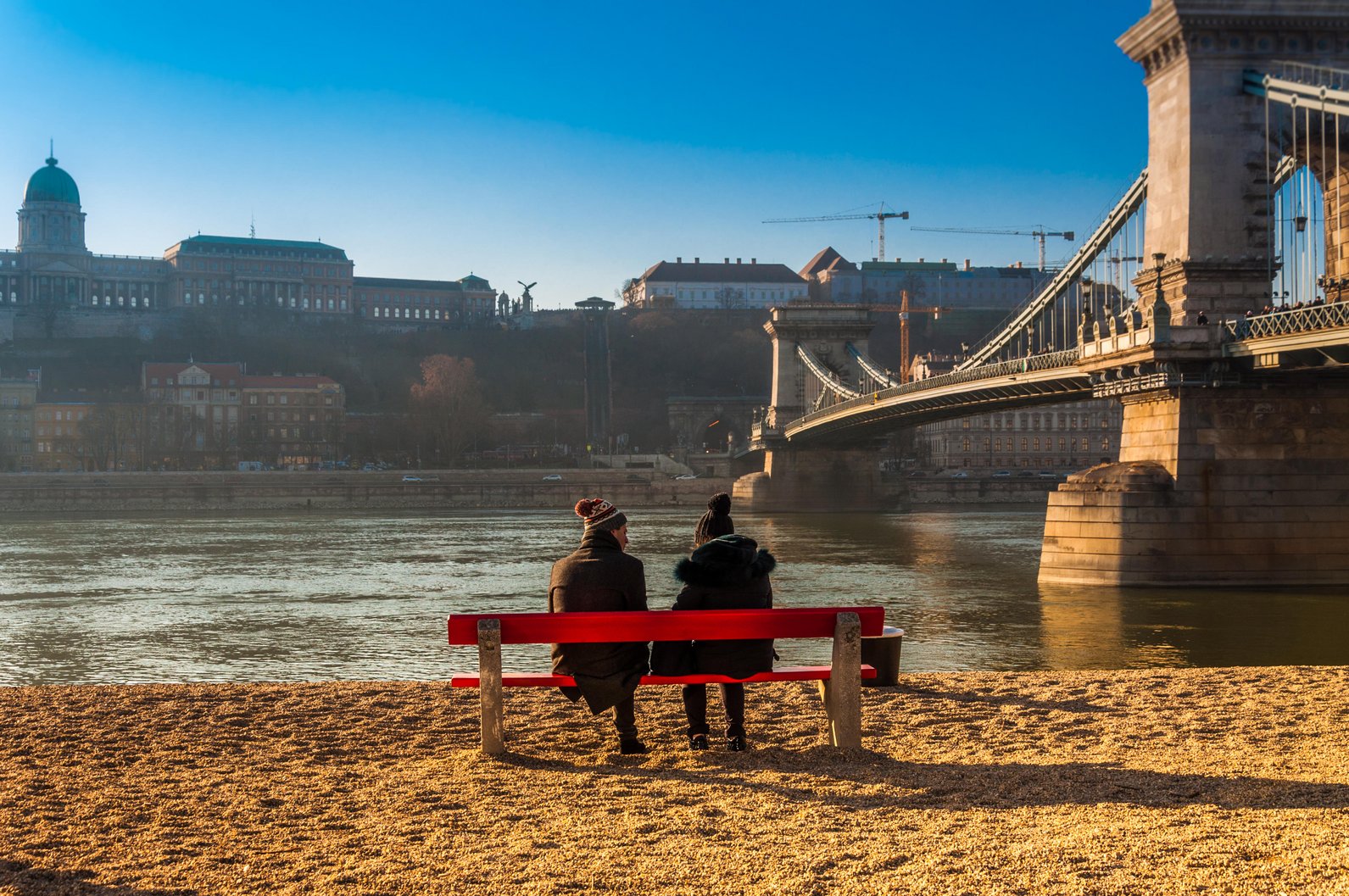 View from behind as two people in dark coats and hats with pom-poms sit on a red bench next to the Danube and the Széchenyi Chain Bridge.