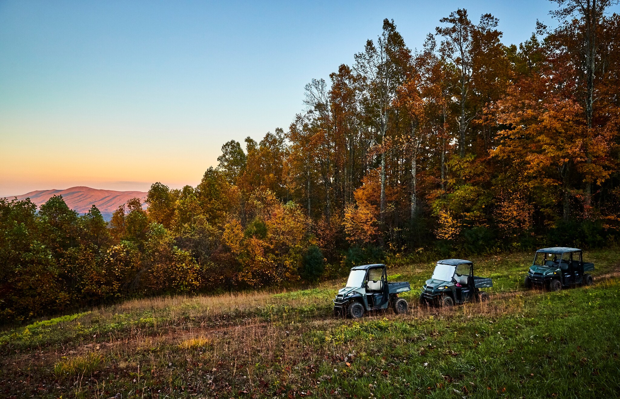 Three small Recreational Terrain Vehicles are parked on a dirt path next to a forest of autumn colors. Mountains are visible in the distance.
