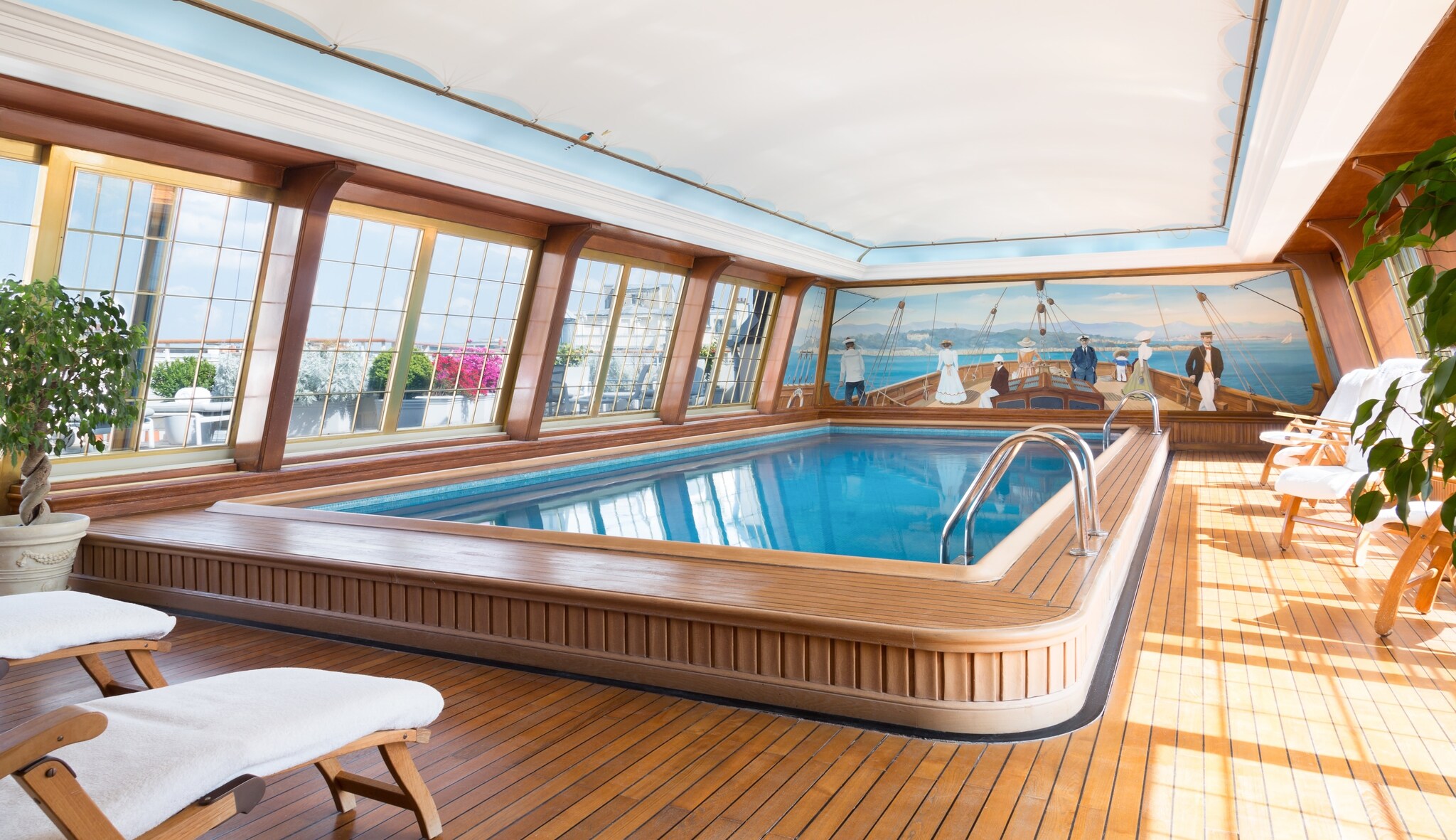 The rooftop pool is enclosed in a bright room with wide windows overlooking the city. The pool itself is slightly raised from the floor.