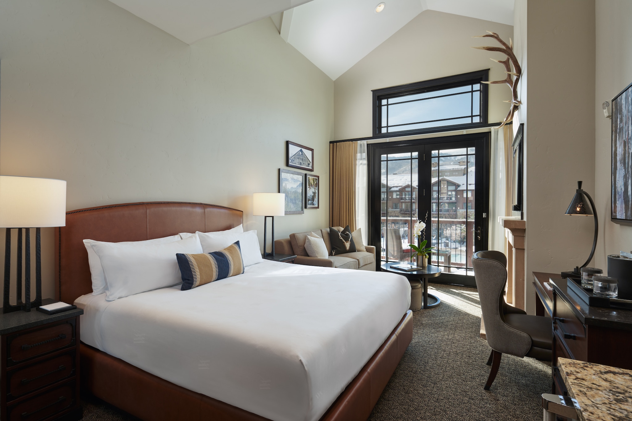 In a King Bedroom Suite, a large bed faces a black desk and wall-mounted stag horns. Glass doors on the far wall open onto a balcony.