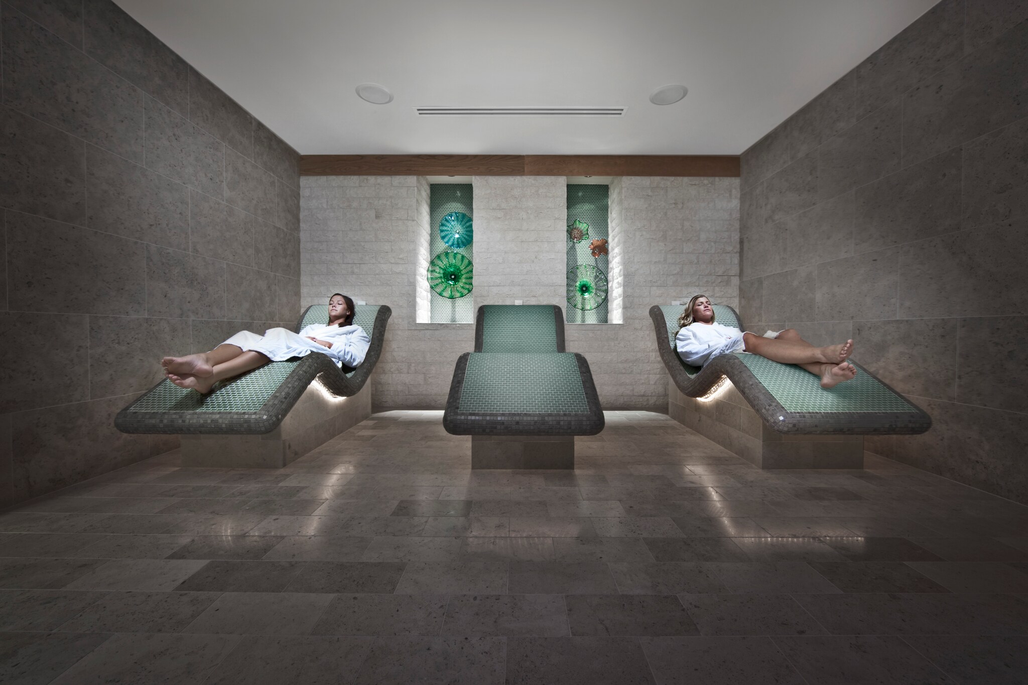 In a gray spa room, an Asian woman and a white woman in white robes lounge on green day beds with a third empty bed between them.