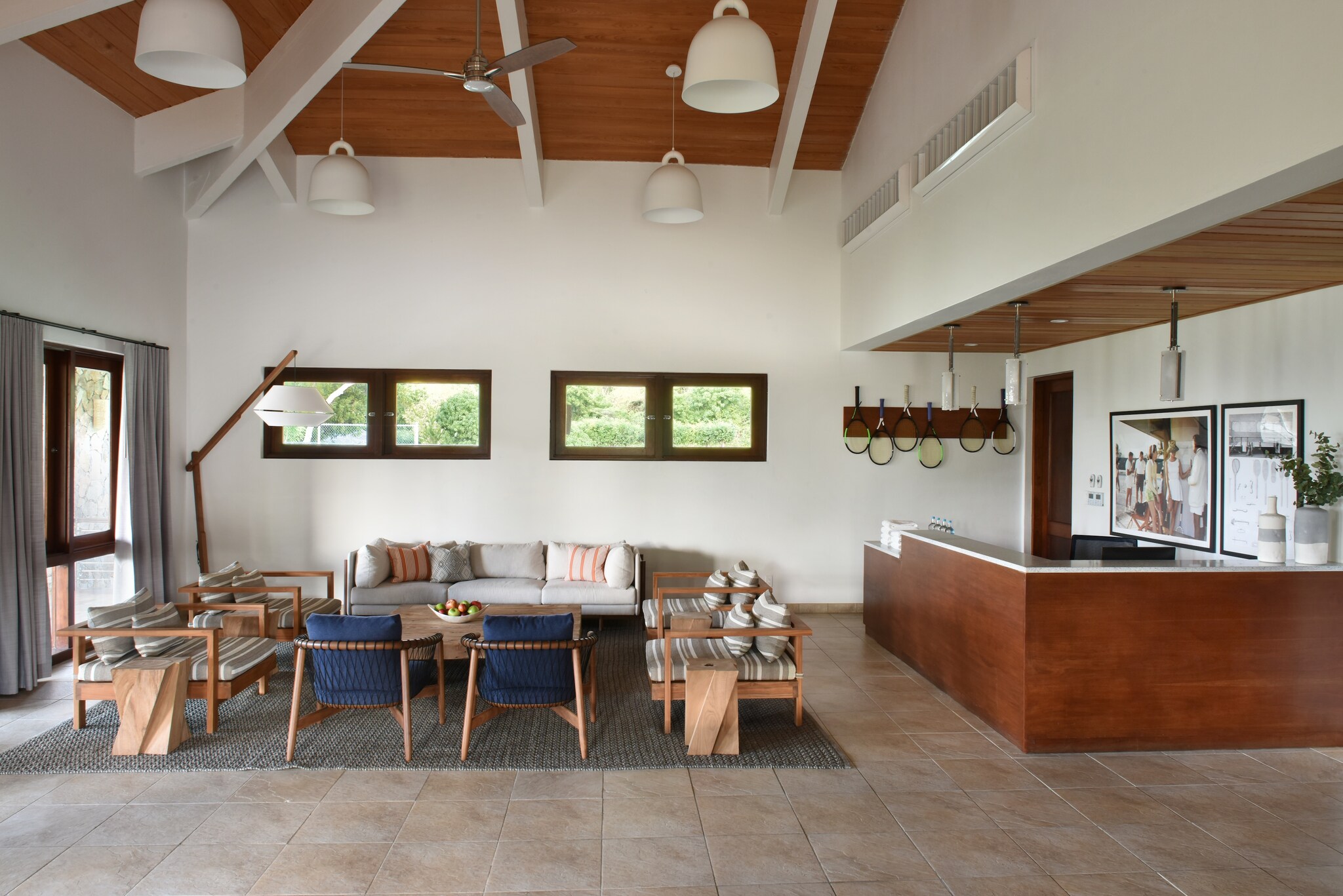 The tennis clubhouse has high ceilings, a seating area with low wooden furniture, and a greeting desk stocked with towels and water bottles.