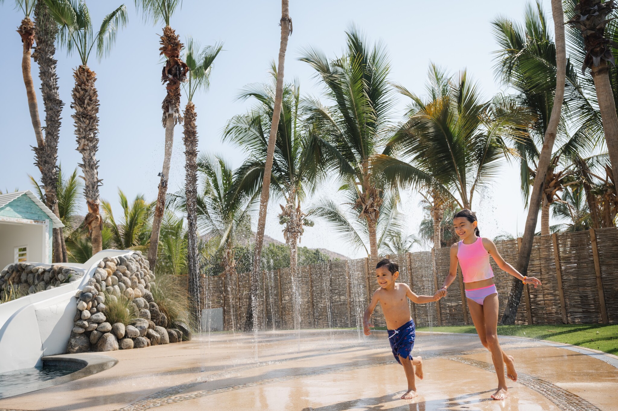 Two Asian children wearing swimsuits run through spurting fountains in the pool complex, the girl holding her younger brother’s hand.