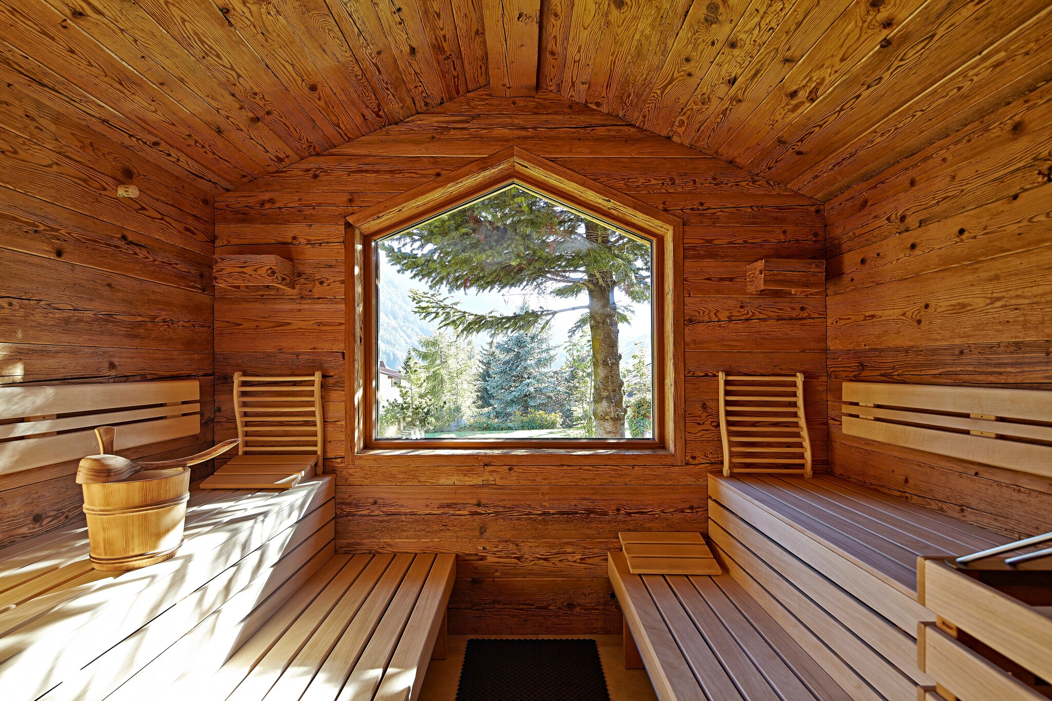 The wooden outdoor sauna has two tiers of wooden benches next to a pentagonal window. A wooden bucket and scoop rests atop the left bench.