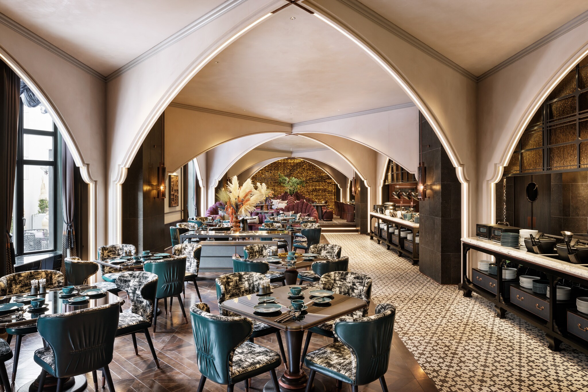 The hotel restaurant has chairs with teal leather backs and patterned seats filling a long dining area next to a tiled serving area.