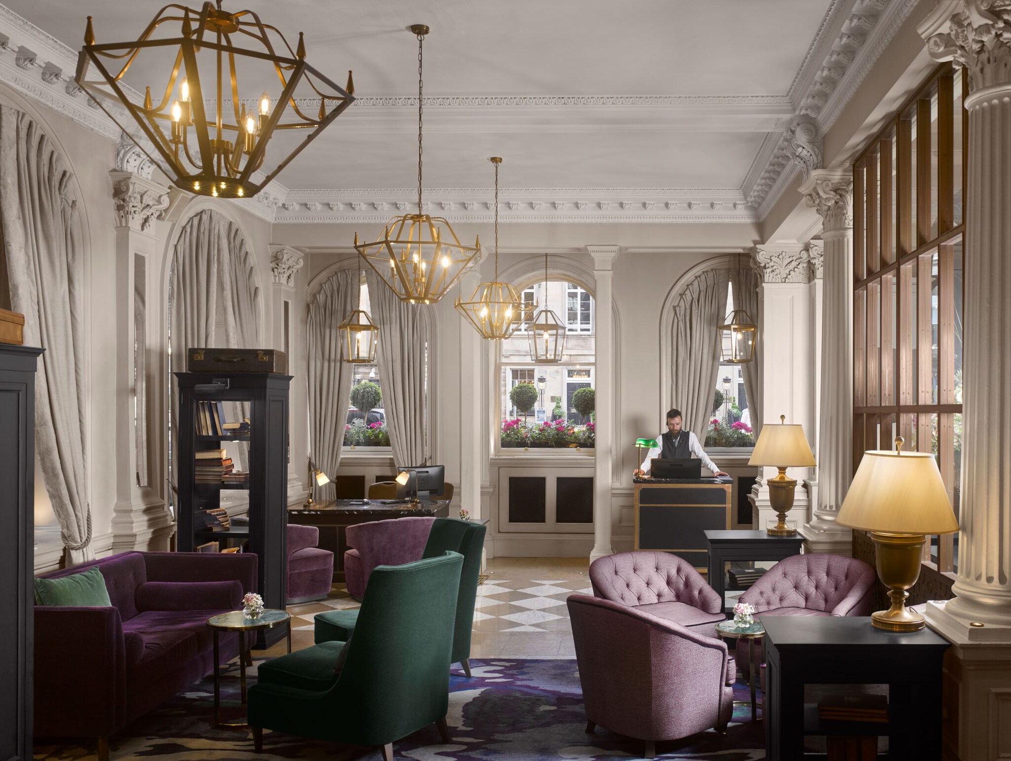 The lobby has checkerboard floors and cream walls. Purple and green armchairs are arranged in clusters between bookshelves.