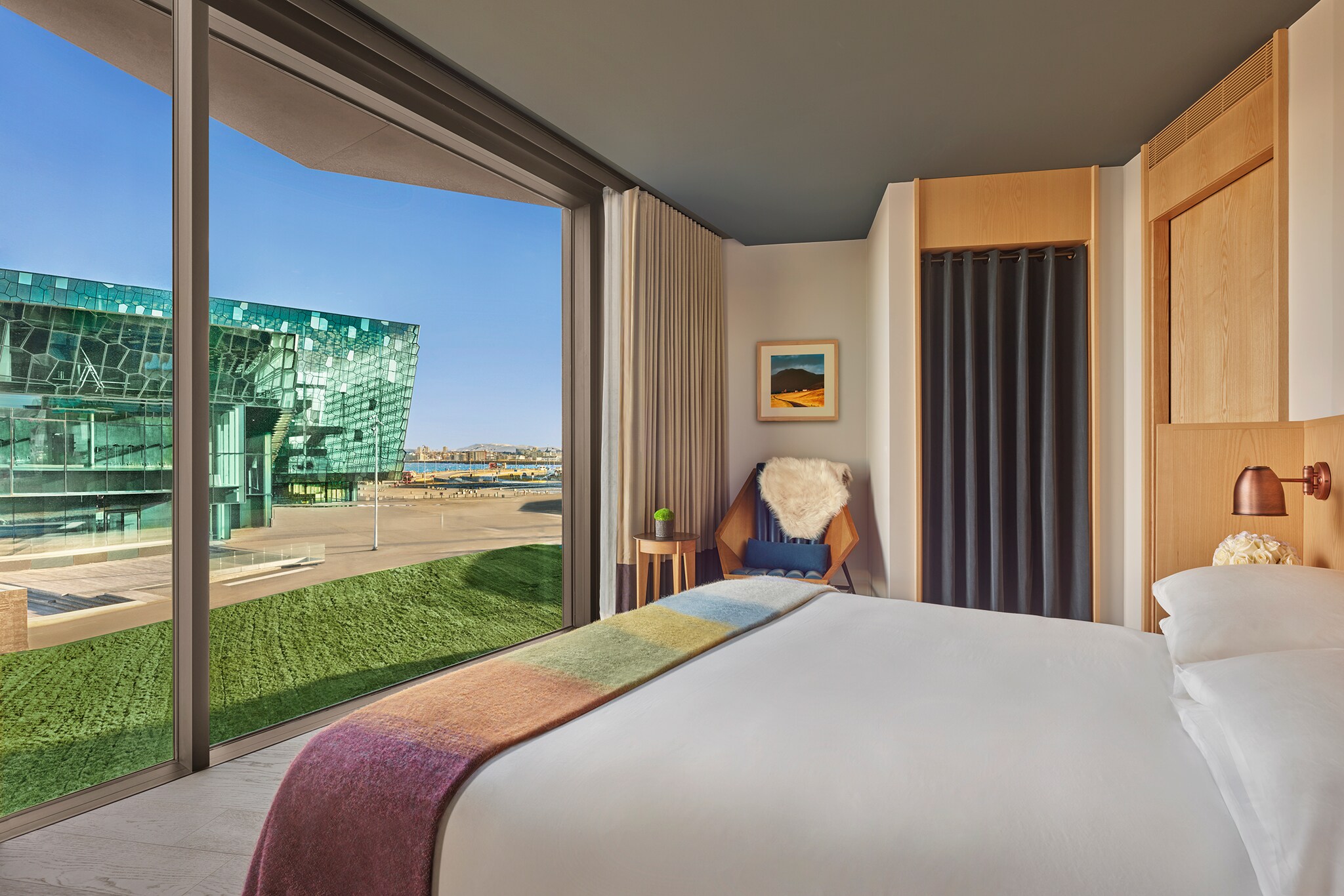 A guest room has a fuzzy rainbow blanket draped around its foot. Floor-to-ceiling windows look past a green lawn at the Harpa concert hall.