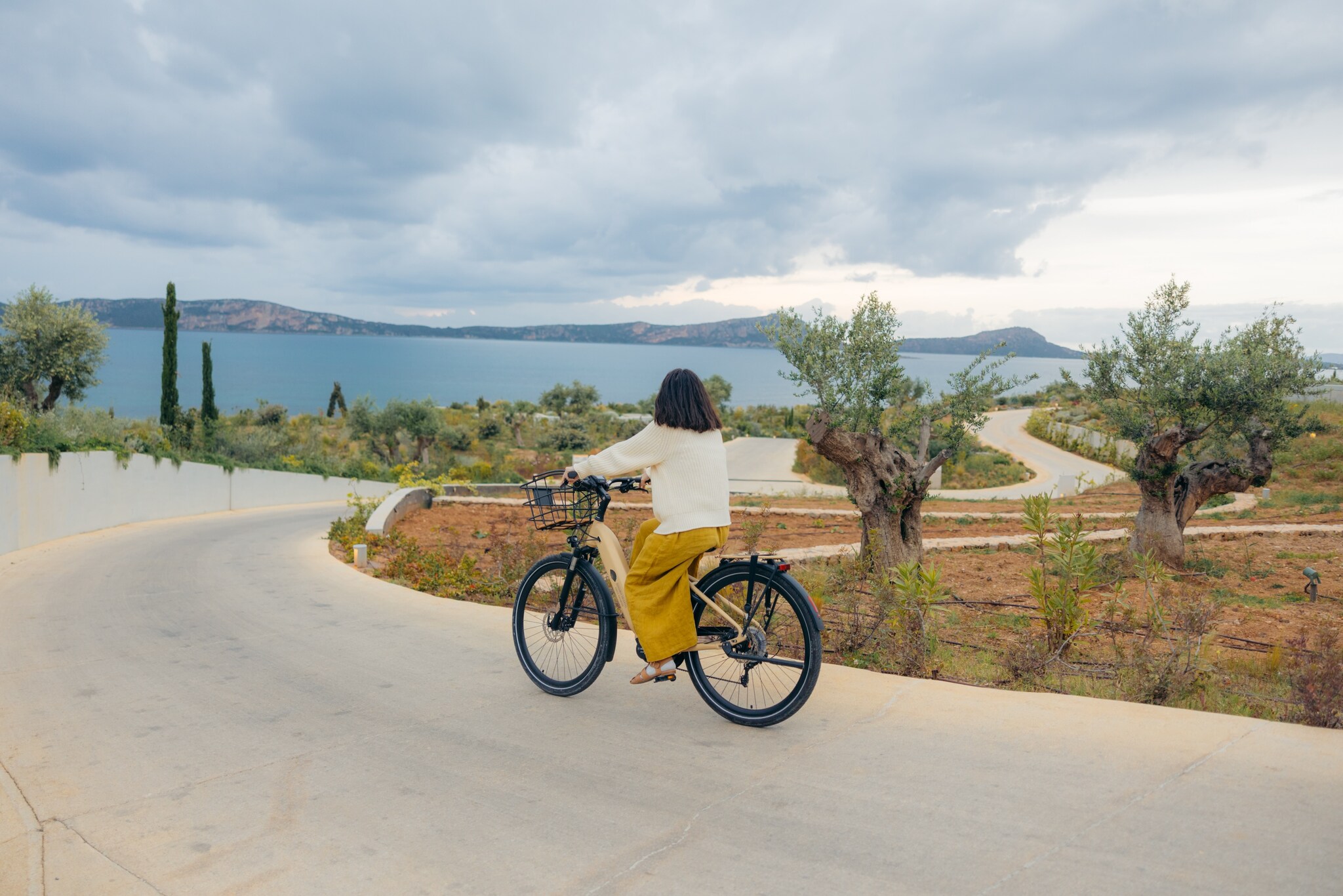 A woman with shoulder-length black hair and mustard-yellow trousers rides a bike down a winding path through the Greek hillside.