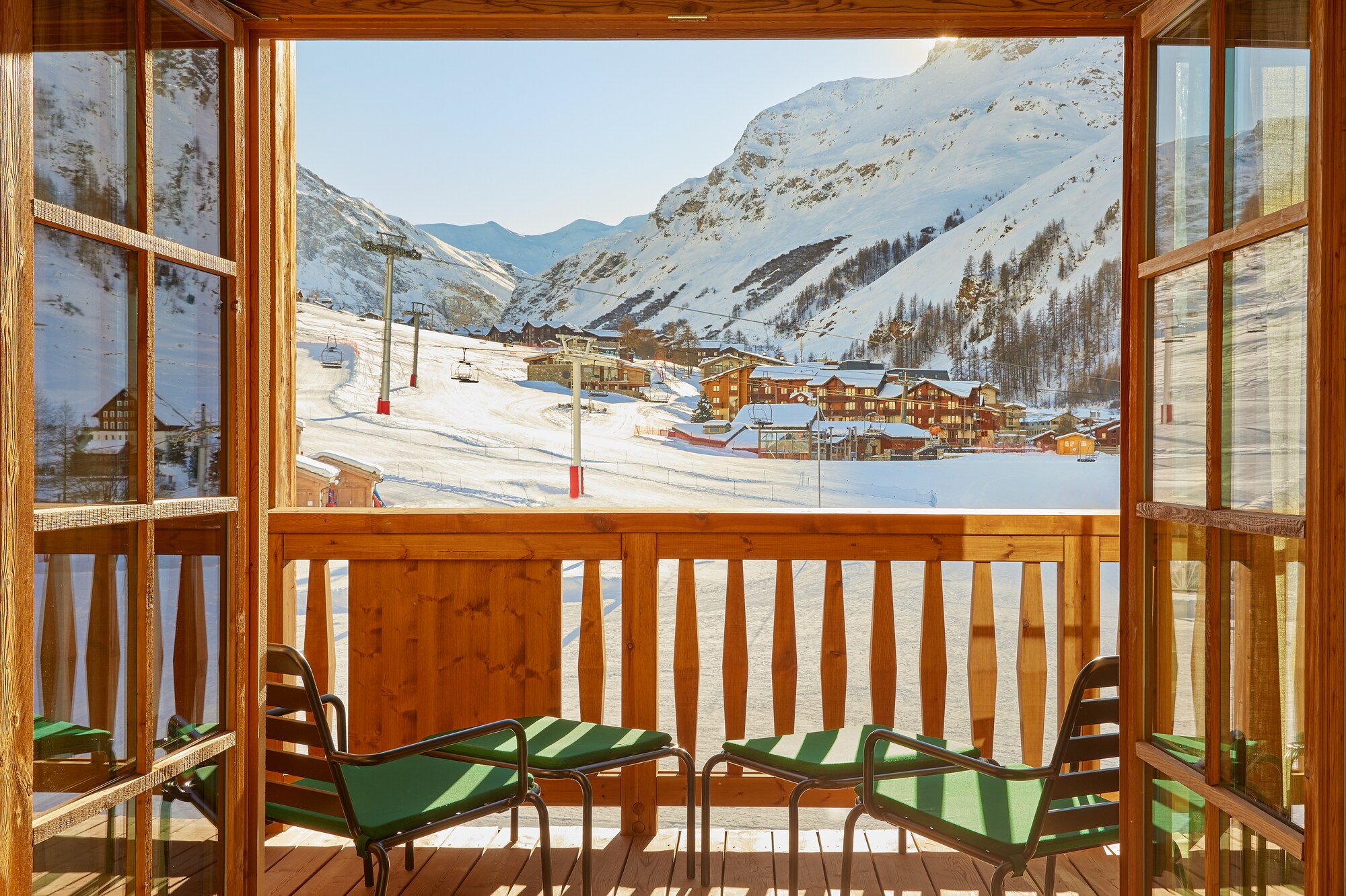 View through two open wooden-framed glass doors over two green lounge chairs and a balcony to a snowy mountainside and village.