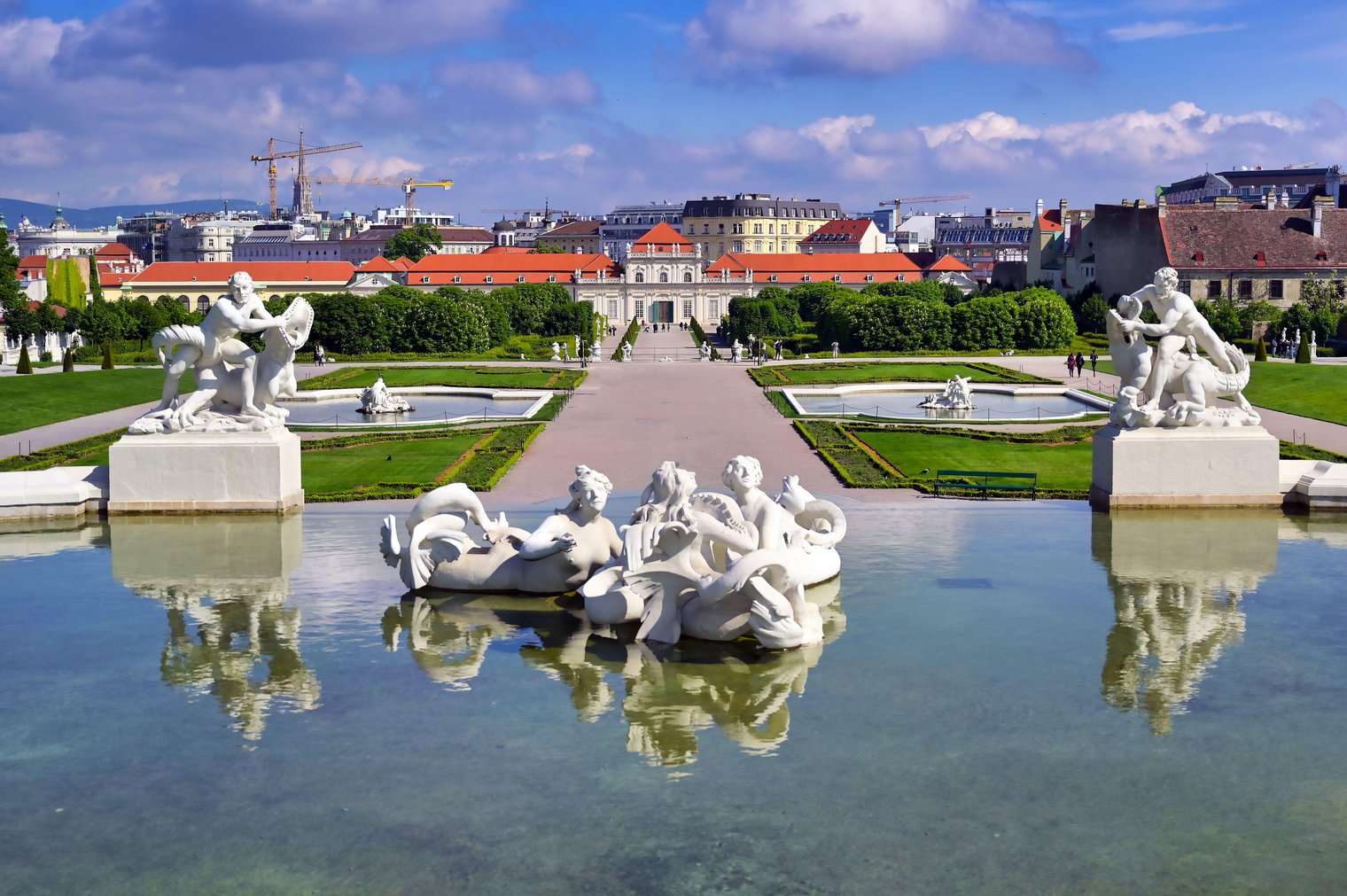 White statues of mermaids entwine in the center of a glassy fountain at the Belvedere Gardens, looking down a wide path towards Lower Belvedere.