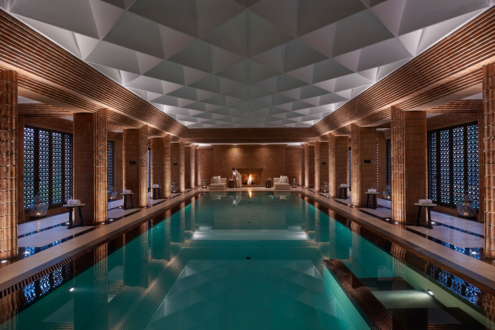 The spa’s indoor pool is long and rectangular. The cavernous room has a geometric white ceiling fixture and striated brown brick columns.