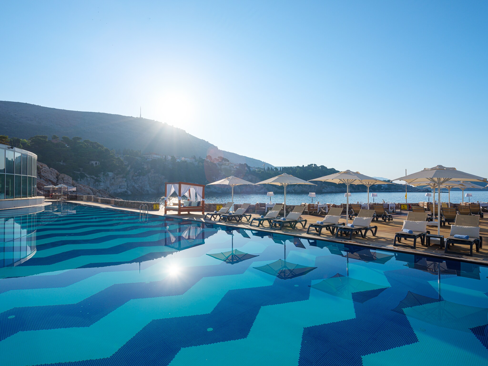 The pool has a zigzag tile pattern along the bottom and the desk is lined with cabanas, white sun beds, and square white umbrellas.
