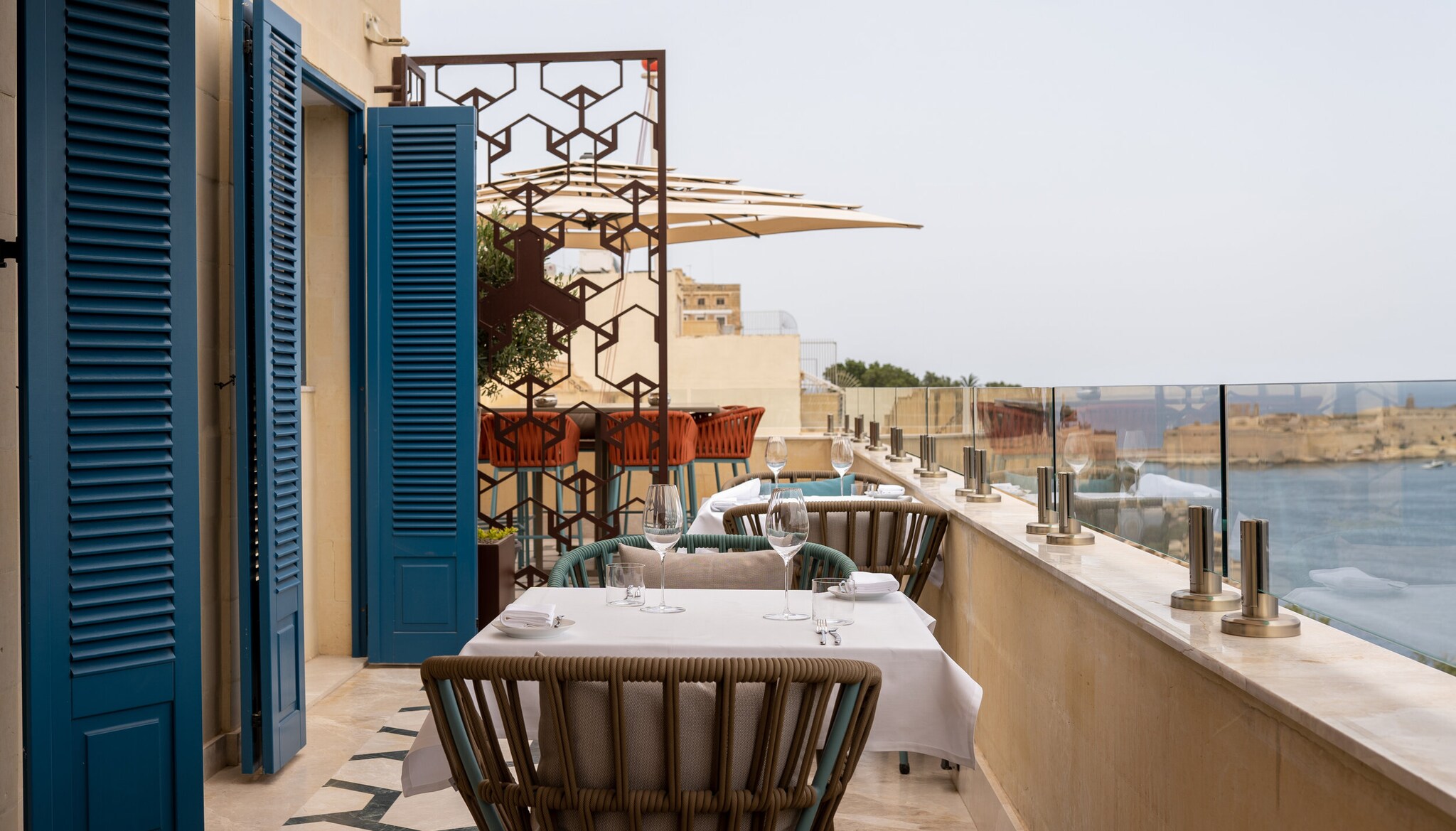 Past blue slatted folding doors, the rooftop terrace dining at ION Harbour features small tables, wicker chairs, and stone railing.