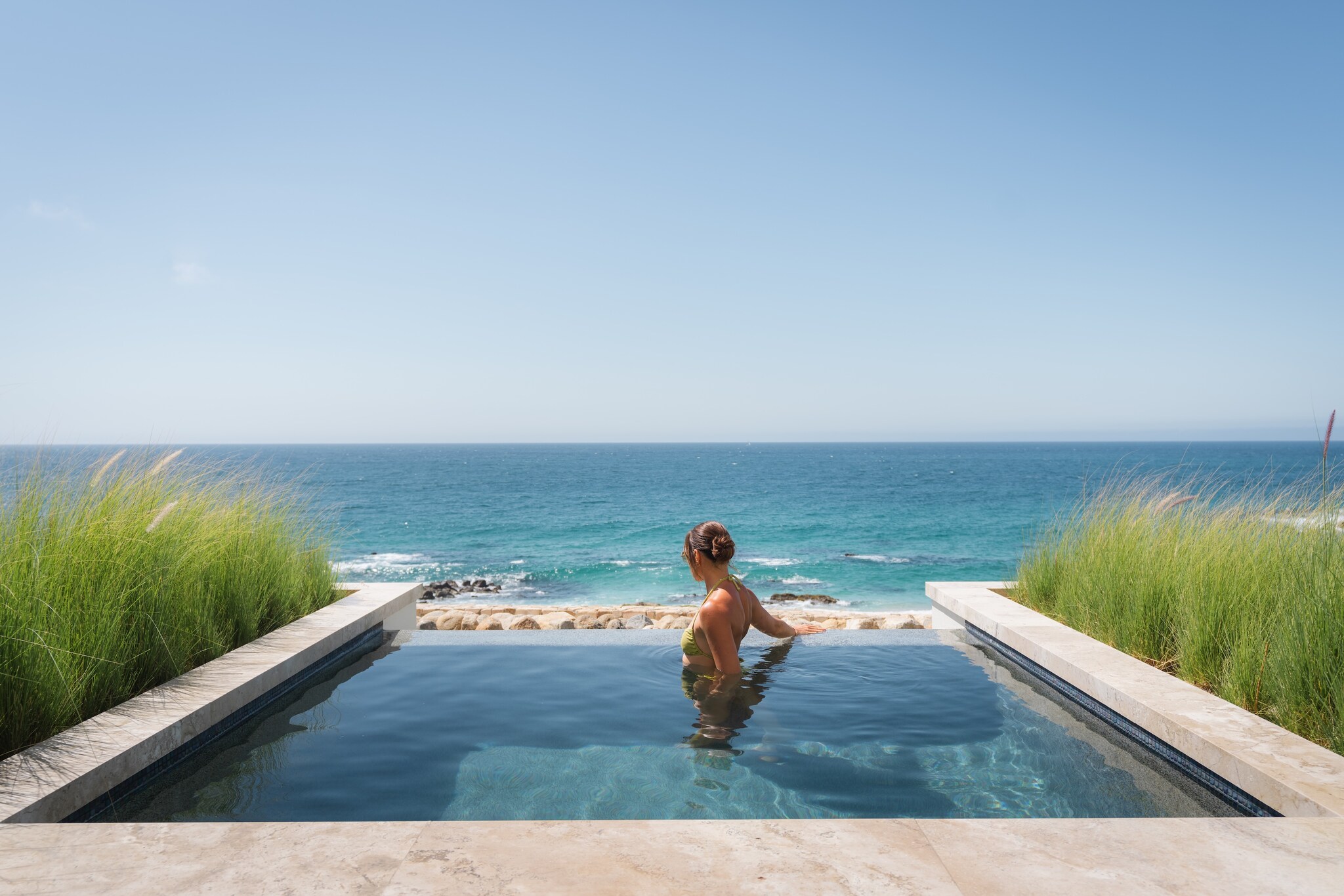 A white woman with her hair in a bun stands in a small infinity pool flanked by beach grass, looking out towards the beach.
