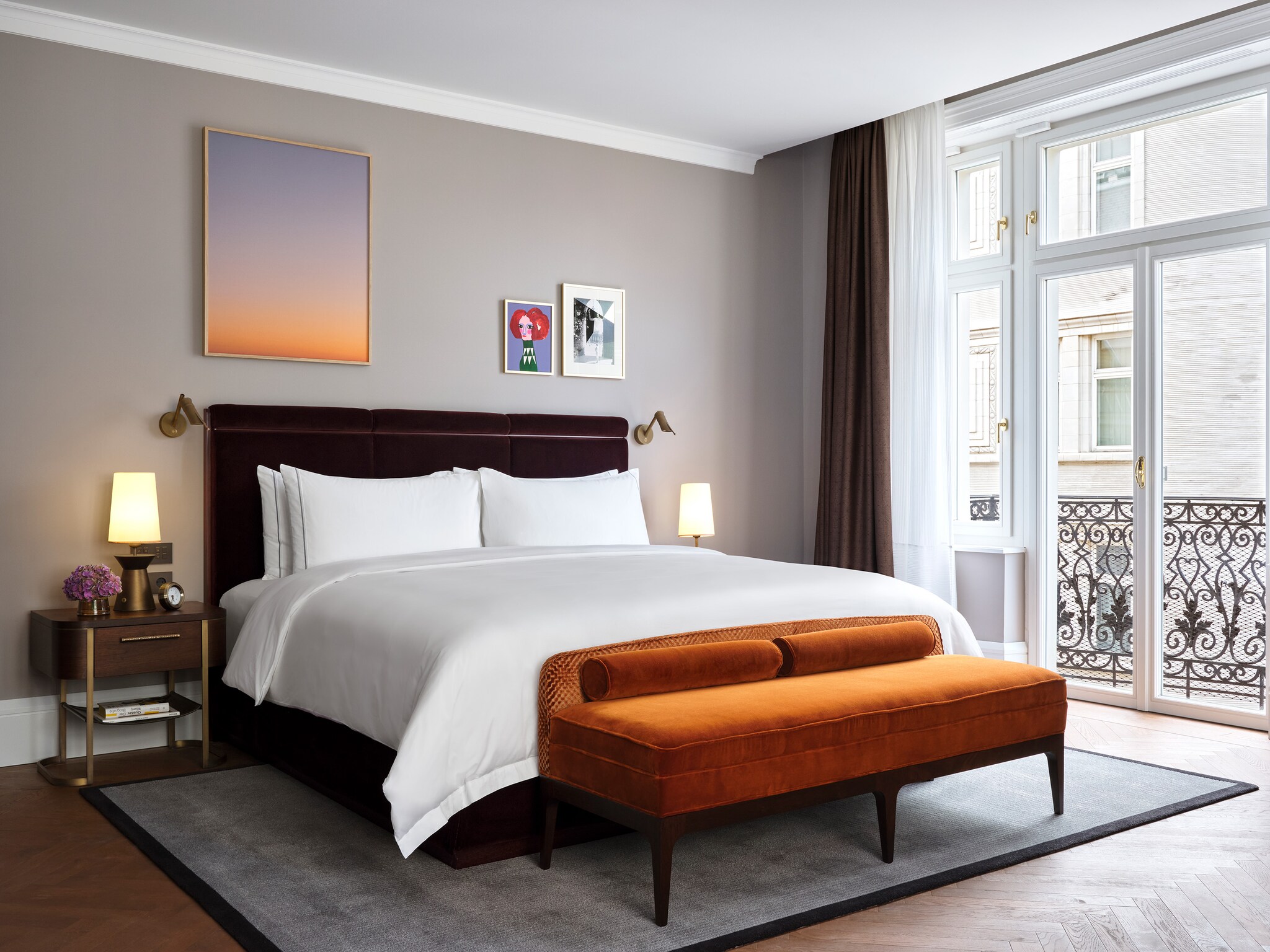 In a Deluxe Room, an orange velvet bench sits at the foot of the bed. White-framed glass doors lead open onto a small balcony.