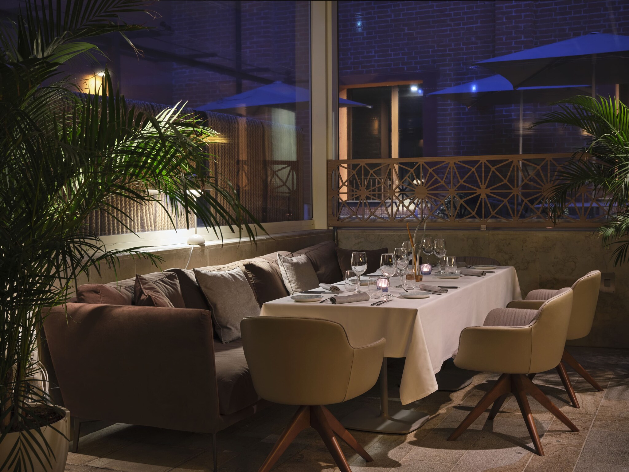 At night, the Osteriva Restaurant is dim, with a couch and several chairs around a long dining table partially shaded by potted palms.