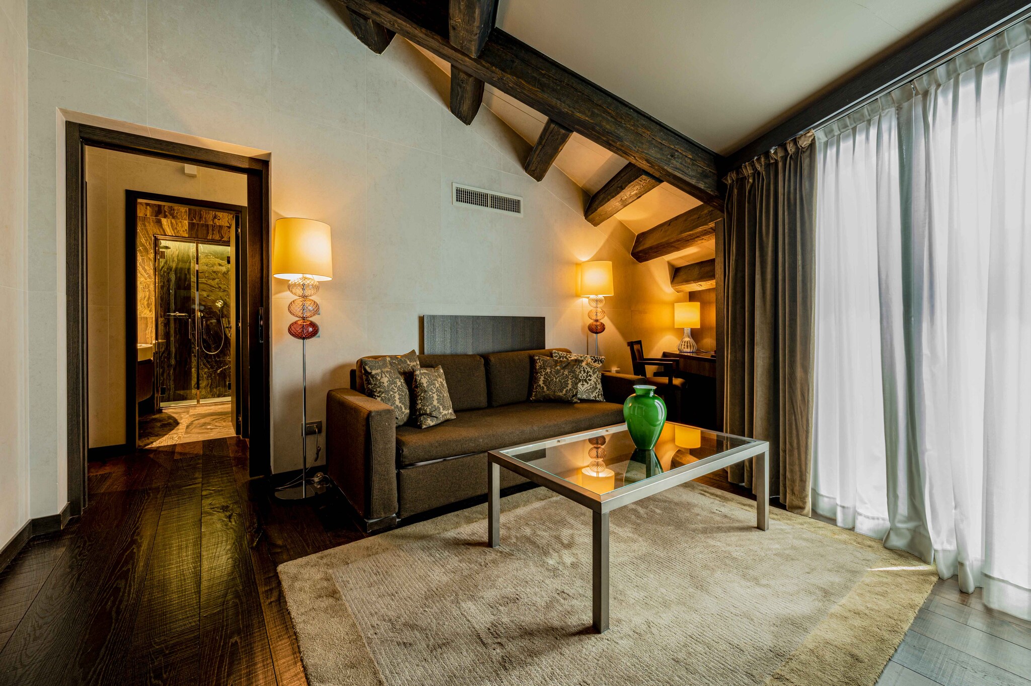 In the living area of the Riva Suite with Canal View, gauzy curtains cover glass doors and a coffee table sits in front of a small couch. 