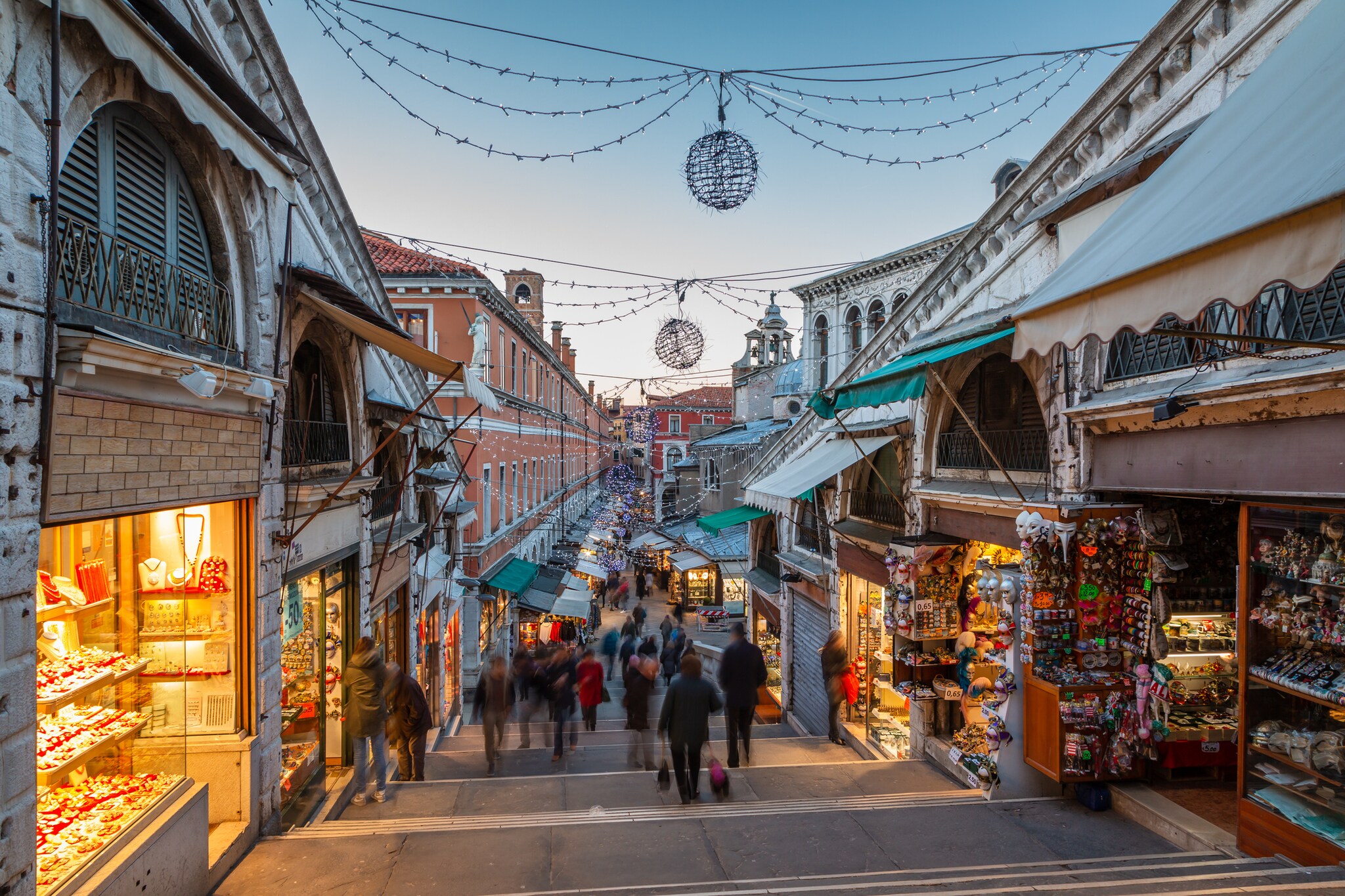 View down wide steps into a Venice street strung with lights and packed with storefronts, including pastry shops and masquerade stores.
