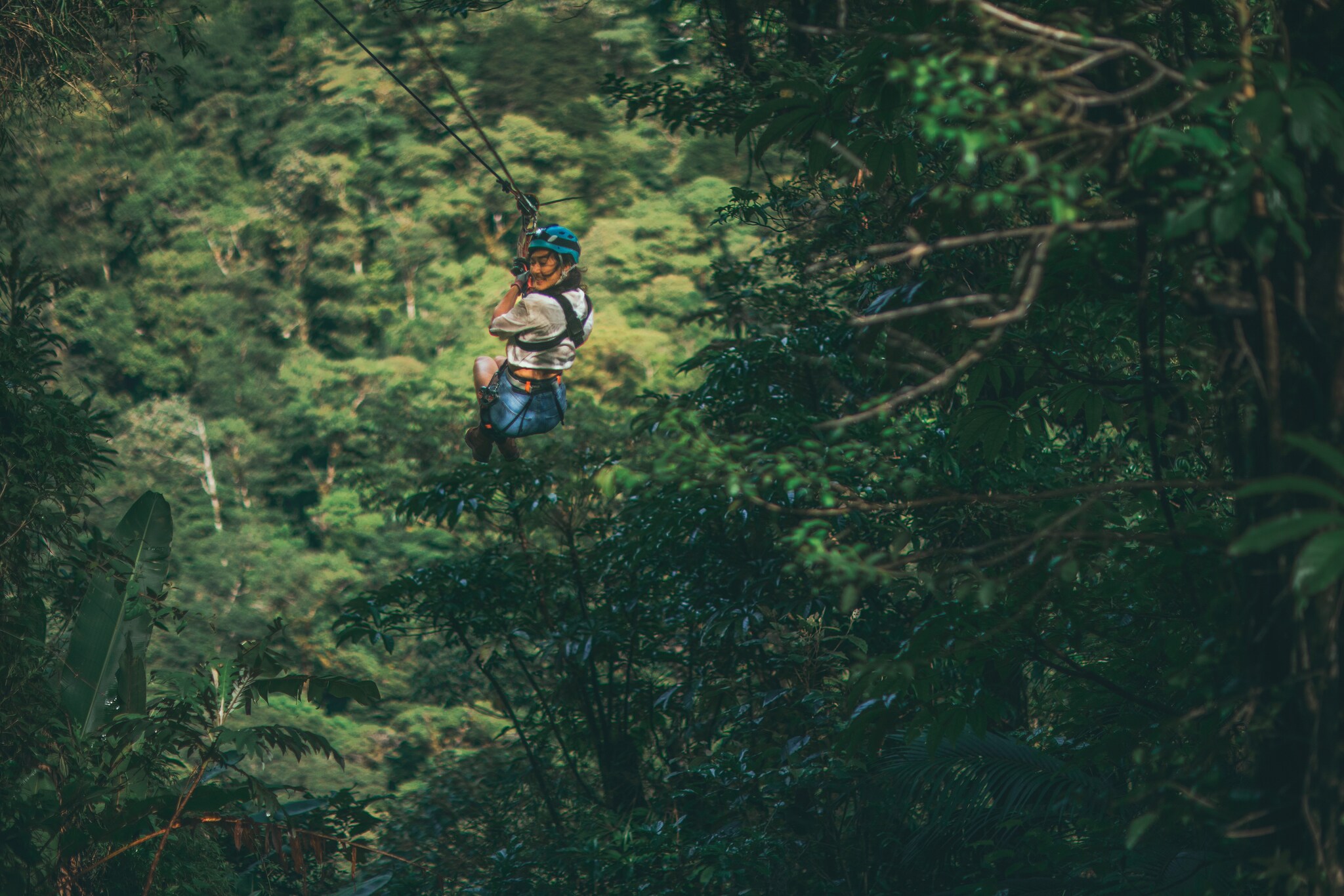 The Asian woman in the blue helmet is partway down a zipline into a jungle, tucking her feet up and looking excitedly back where she came.