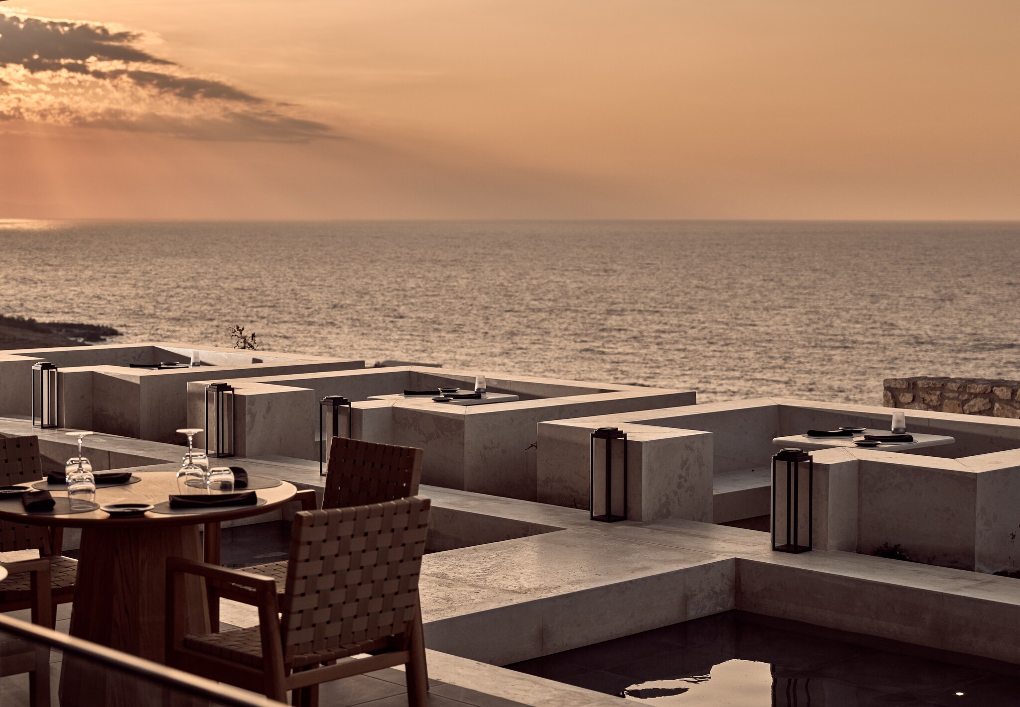 At sunset, the angular white stone booths and wooden tables of Cretamos Restaurant are washed in pale orange light glancing off the sea.