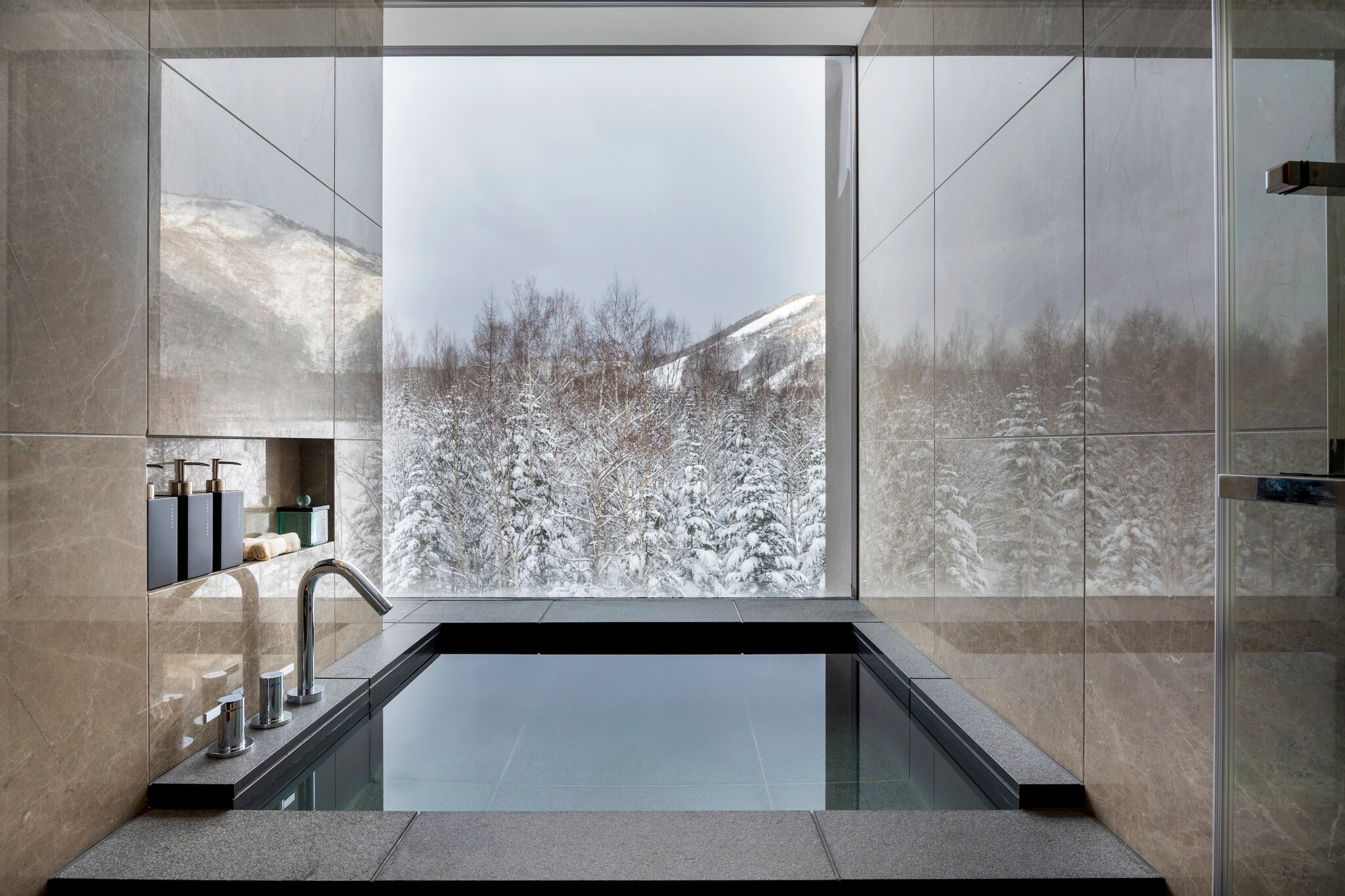 A guest room bathroom has a deep, square soaking tub next to a window overlooking snowy trees.