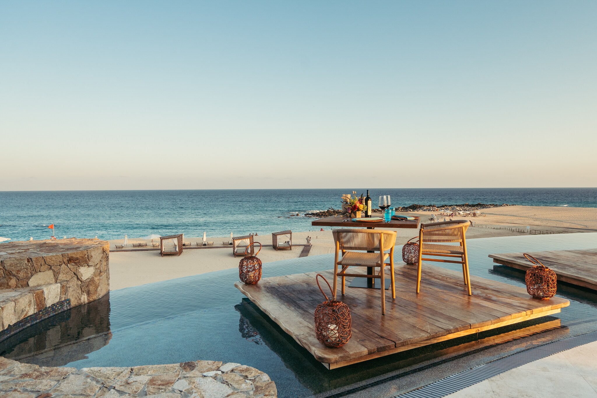 A square wooden platform in a small pool overlooking the beach is set with wine and plates for a romantic dinner for two.