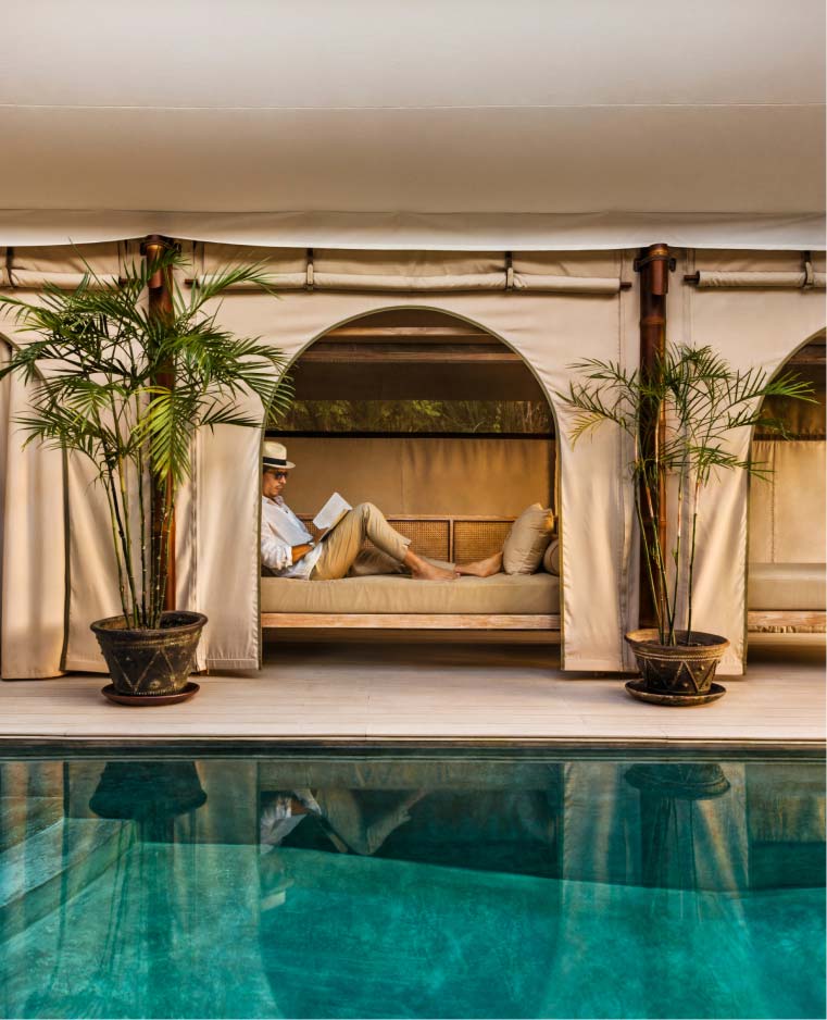 A middle-aged Latino man reads and reclines in a cabana shaded by a beige tent and flanked by potted palm trees.