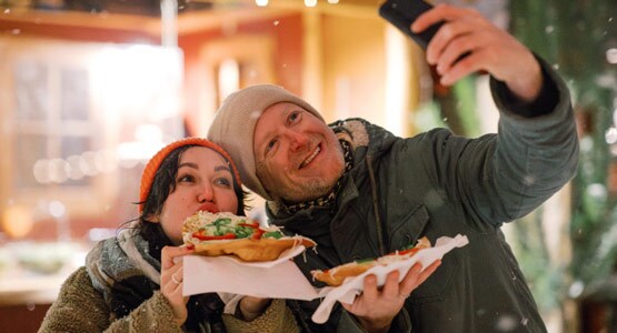 A pale middle-aged man and younger woman pose for a selfie. They’re dressed in parkas with snowflakes swirling around, and eating flatbread.
							