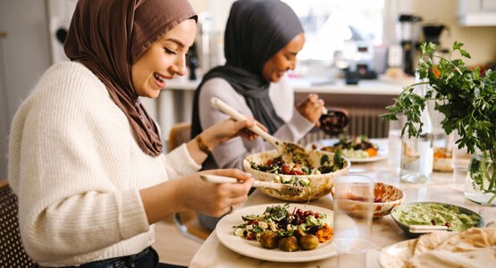 Two young women wearing hijabs laugh as they sit at a dinner table serving themselves falafel and salad