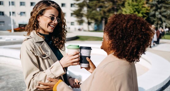 Two women, one with dark skin and one with medium-light skin, smile and toast with their reusable coffee cups as they meet on the street