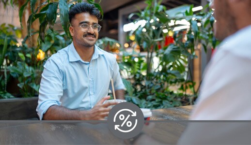A South Asian man with glasses and a button-down shirt sits across a table from an out-of-focus person, both drinking coffee.