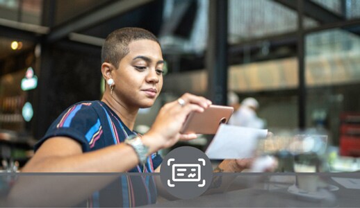 A Black person with close-cropped hair and a striped T-shirt photographs a check with their phone while sitting at a café.