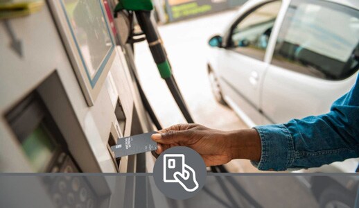 A Black man’s hand inserts an American Express Debit Card into a gas pump, a silver sedan out-of-focus in the background.