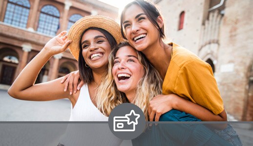 Two white women and a Black woman, all dressed colorfully, crowd close and smile wide as they pose for a picture on a sunny street.