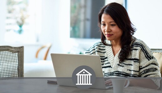 An Asian woman in a white striped sweater sits at a table in her home with a cup of coffee, adding funds on her laptop.