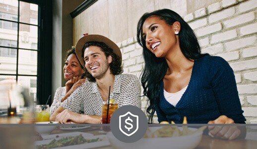 Two young Black women and a young white man, all dressed casually, sit side by side enjoying salad, fries, and iced tea in a restaurant.