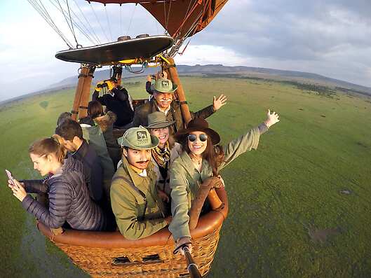 A group of vacationers and their guides spread their arms joyfully in a hot air balloon over the Maasai Mara