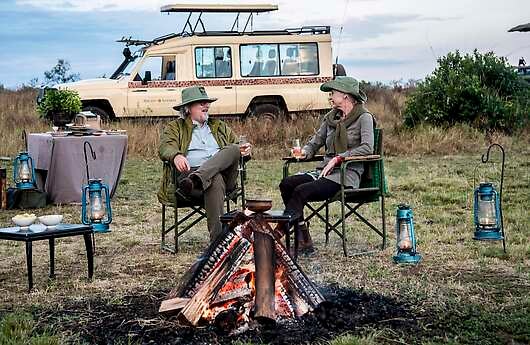 Two travelers converse in camp chairs next to a fire while a safari truck idles nearby