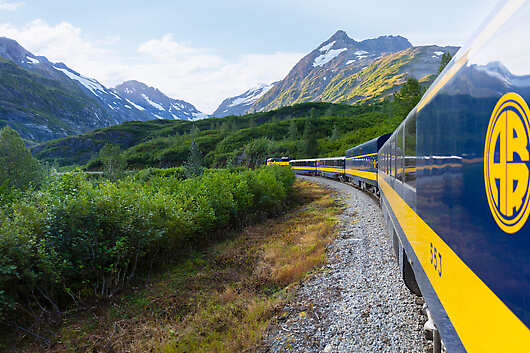 An Alaska Railroad train winds its way through a lush valley between snow-dappled mountains
