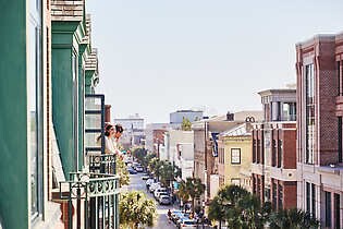 An adult female and adult male on a guestroom balcony at The Charleston Place hotel overlooking a street in downtown Charleston, South Carolina.