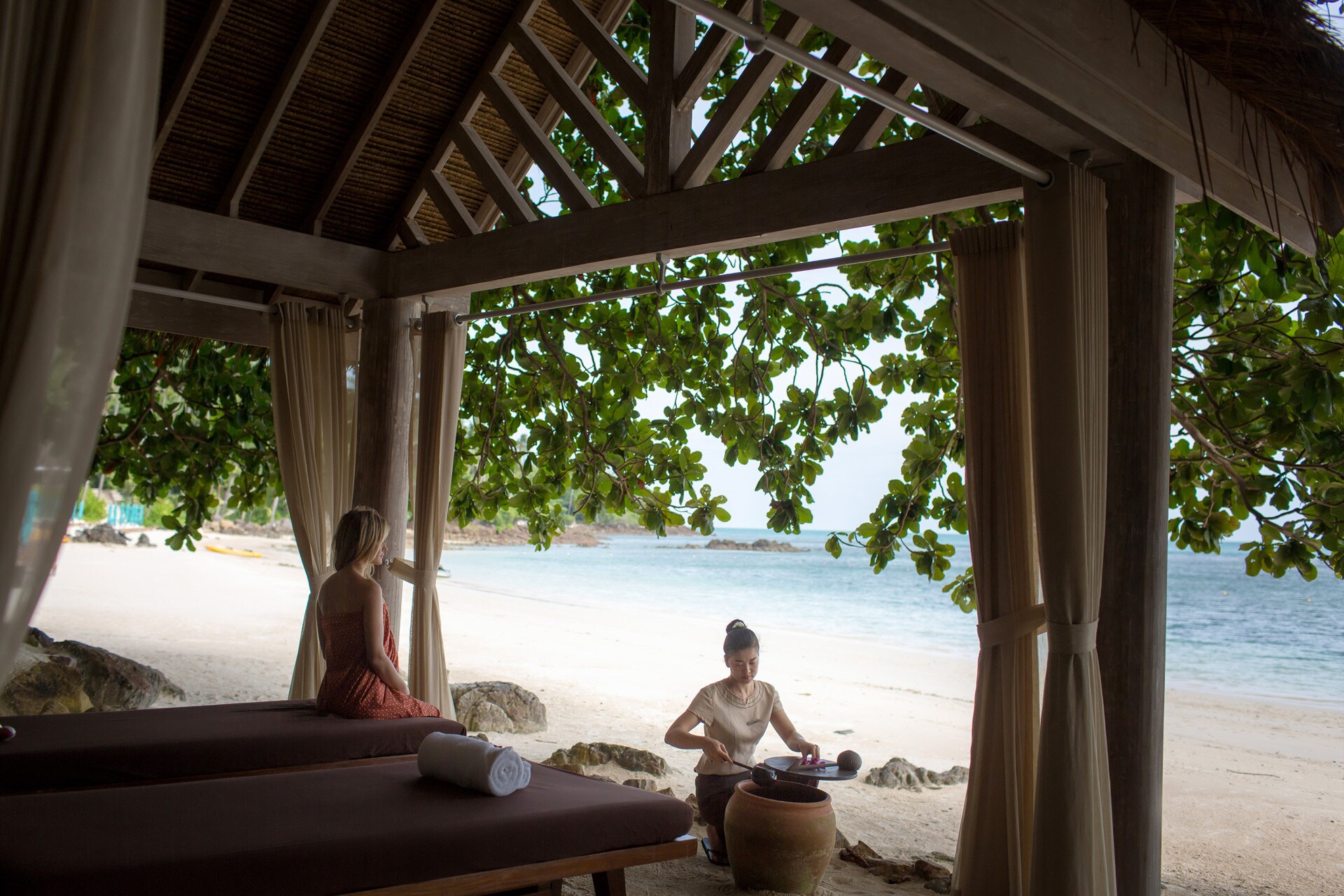 In a beachside gazebo, a white woman in an orange wrap sits on a massage bed while a female employee ladles liquid from a jug.