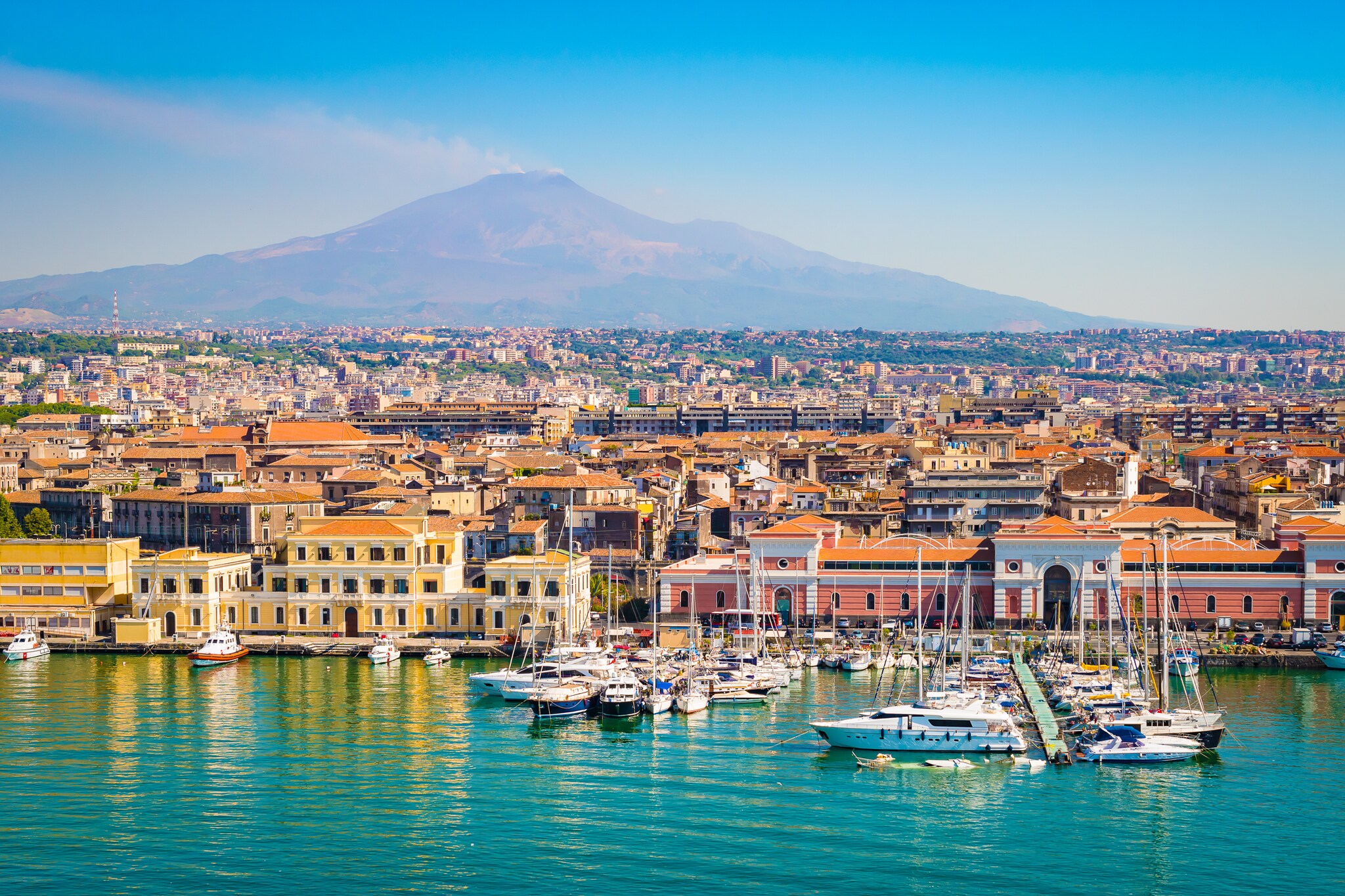 Aerial view of Catania harbor. Boats line the marina in front of brightly colored pastel buildings, most with terracotta roofs.