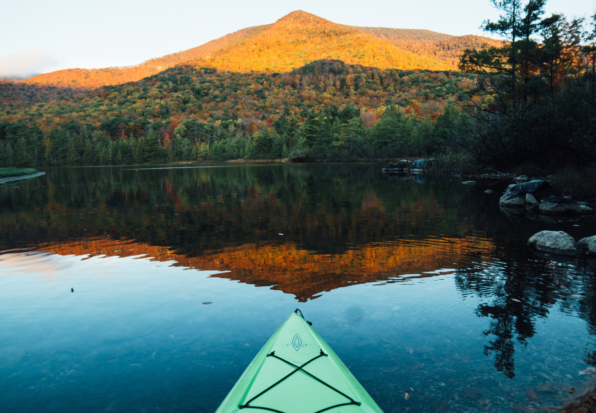 View across the bow of a seafoam green kayak gliding on a glassy lake, which reflects a mountain covered in trees turning red and green.