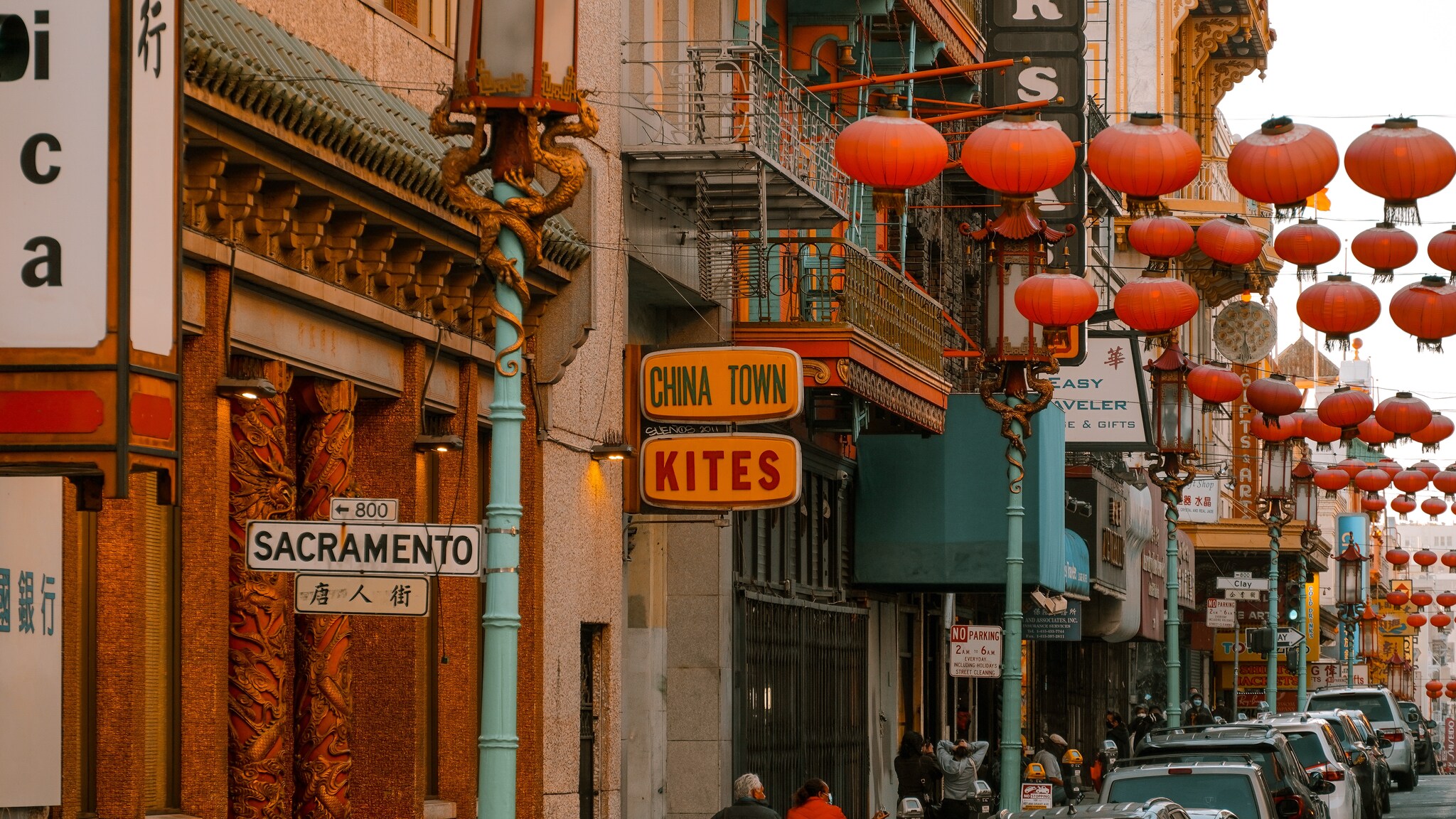 A sign for Sacramento Street is posted on a road crowded by shop signs under lines of red lanterns and street lamps with bronze dragons.