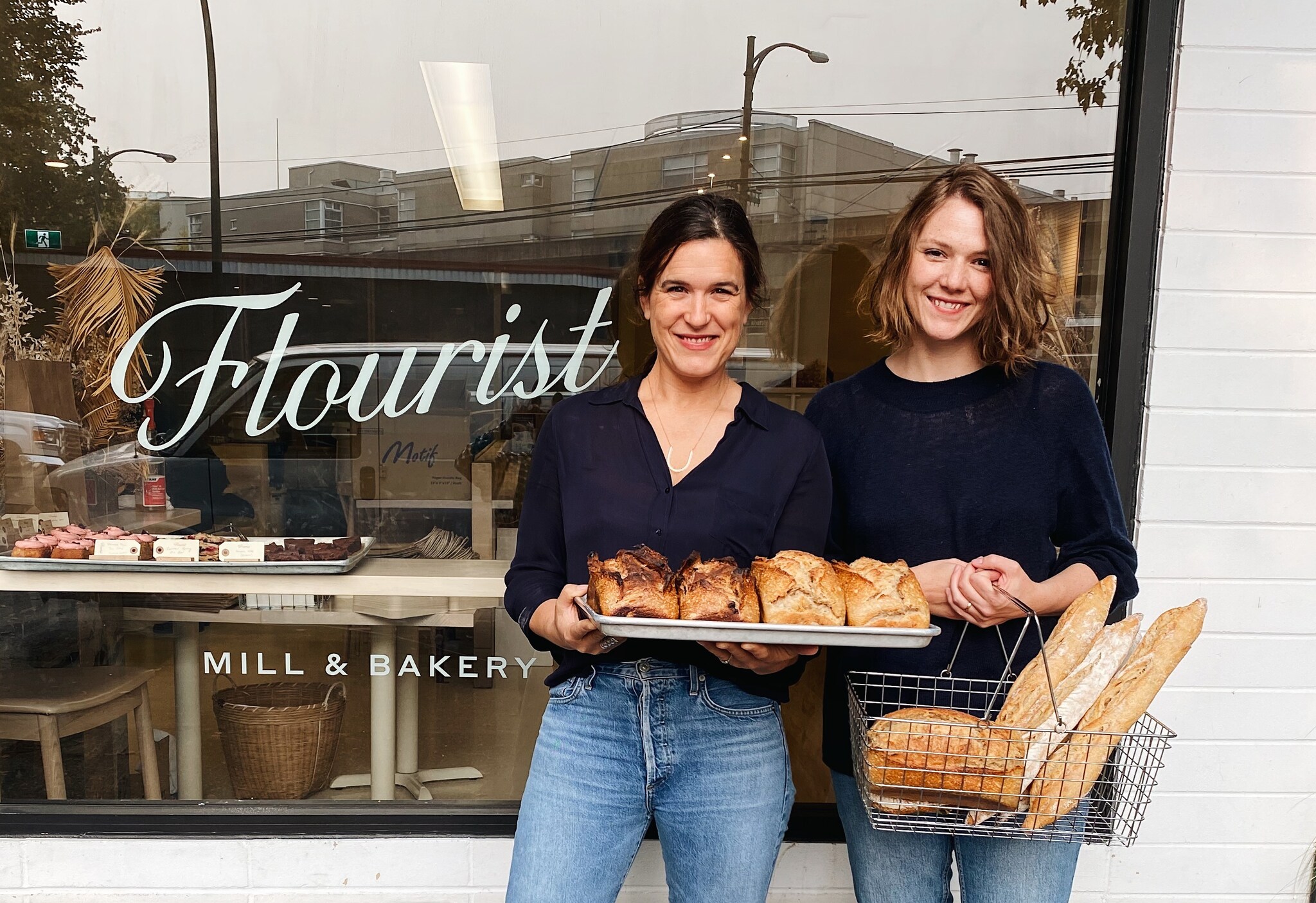 Two young white women pose smiling with loaves of bread beside a window etched with the store name: “Flourist.”