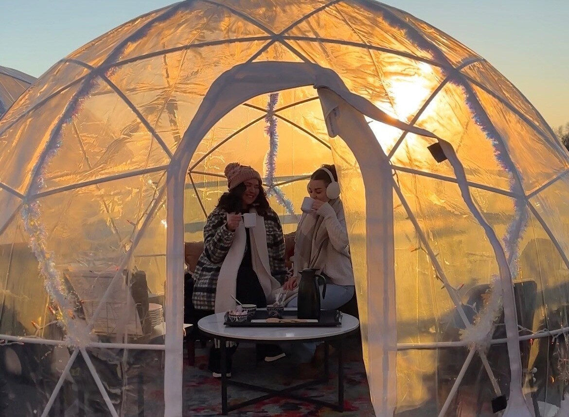 Two women dressed for cold weather sip hot drinks inside a plastic geodesic dome at sunset.