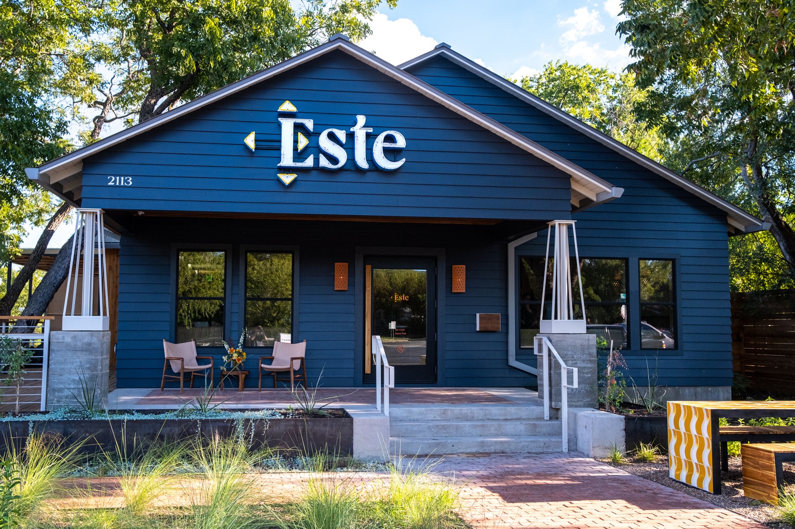 The blue exterior of Este features a bench with chairs, a couple of chairs on the porch, and compass points around the E of the name.