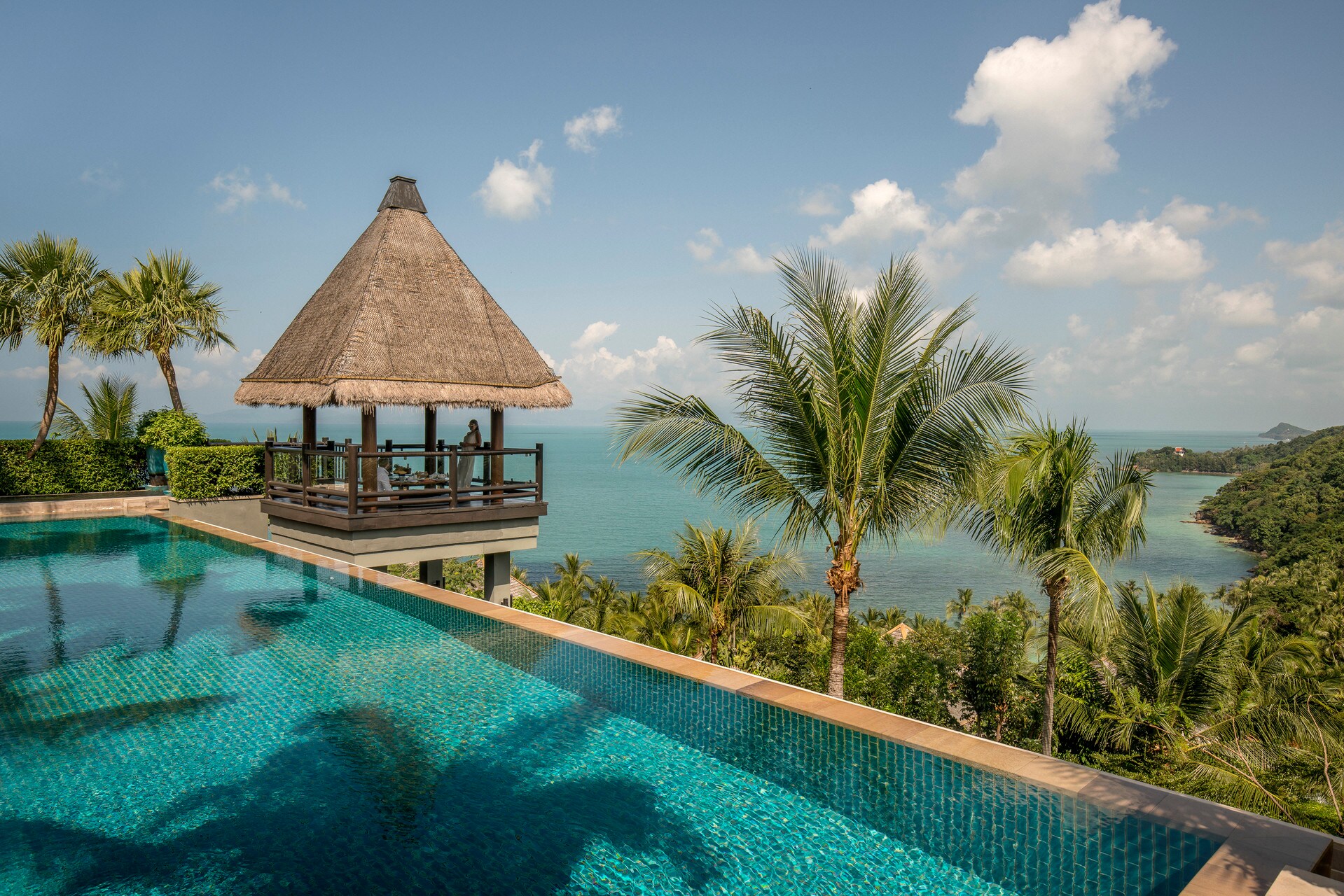 A cabana with a steep thatched roof perches between a blue-tiled pool and a palm-covered hillside leading down to the sea.